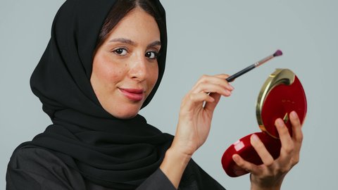 مفهوم العناية بالجمال والبشرة، الترويج لمنتجات و مساحيق التجميل، الاهتمام بالمظهر الخارجي, استخدام المستحضرات التجميلية, بورتريه مقرب لامرأة عربية خليجية سعودية محجبة ترتدي العباءة السوداء تستعمل فرشاة ظل العيون تقوم بوضع الميكب, خلفية رمادي