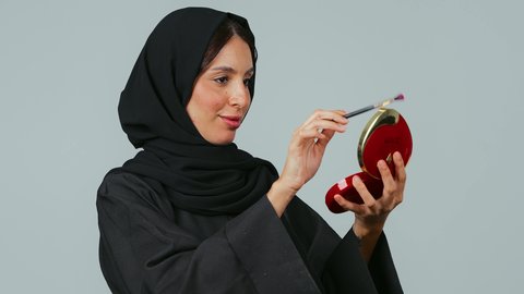 الترويج لمنتجات و مساحيق التجميل، الاهتمام بالمظهر الخارجي, استخدام المستحضرات التجميلية, مفهوم العناية بالجمال والبشرة, بورتريه مقرب لامرأة عربية خليجية سعودية محجبة ترتدي العباءة السوداء تستعمل فرشاة ظل العيون تقوم بوضع الميكب, خلفية رمادي