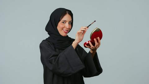 الاهتمام بالمظهر الخارجي, استخدام المستحضرات التجميلية, الترويج لمنتجات و مساحيق التجميل, مفهوم العناية بالجمال والبشرة, بورتريه لامرأة عربية خليجية سعودية محجبة ترتدي العباءة السوداء تستعمل فرشاة ظل العيون تقوم بوضع الميكب, خلفية رمادي