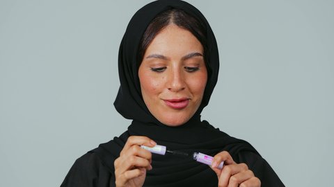 Using mascara to highlight the beauty of the eyes, educational courses for makeup tutorials, attention to beauty and appearance, a portrait of a Saudi Arabian Gulf woman wearing a hijab and abaya applying makeup, gray background.
