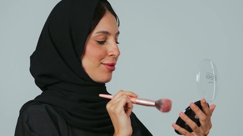 The concept of adornment and beautification, marketing of cosmetics and beauty products, care for the external appearance, use of makeup, a close-up portrait of a Saudi Gulf Arab woman wearing a hijab and a black abaya using a blush brush, gray background.