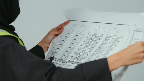 العمل في القطاع الهندسي, تدقيق المخططات والتصاميم المعمارية, بورتريه  لمهندسة عربية خليجية سعودية ترتدي الحجاب والعباءة السوداء وسترة الحماية تمسك مخطط هندسي , إدارة المشاريع الهندسية, مهن ووظائف نسائية,  خلفية رمادية