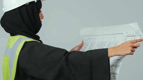 إدارة المشاريع الهندسية,  تدقيق المخططات والتصاميم المعمارية, بورتريه  لمهندسة عربية خليجية سعودية ترتدي الحجاب والعباءة السوداء وسترة الحماية تمسك مخطط هندسي ,مهن ووظائف نسائية, العمل في القطاع الهندسي, خلفية رمادية