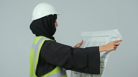 تدقيق المخططات والتصاميم المعمارية, بورتريه  لمهندسة عربية خليجية سعودية ترتدي الحجاب والعباءة السوداء وسترة الحماية تمسك مخطط هندسي , إدارة المشاريع الهندسية, مهن ووظائف نسائية, العمل في القطاع الهندسي, خلفية رمادية