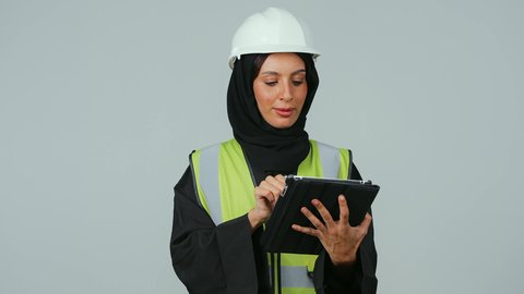 بورتريه  لمهندسة عربية خليجية سعودية ترتدي الحجاب والعباءة السوداء وسترة الحماية تمسك بيدها جهاز التابلت ، التطور والتقدم التكنولوجي في مجال الصناعة والهندسة، المتابعة والإشراف على العمل عبر الجهاز اللوحي ,خلفية رمادية