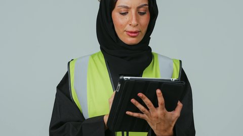 التطور والتقدم التكنولوجي في مجال الصناعة والهندسة، المتابعة والإشراف على العمل عبر الجهاز اللوحي , بورتريه  لمهندسة عربية خليجية سعودية ترتدي الحجاب والعباءة السوداء وسترة الحماية تمسك بيدها جهاز التابلت ،  خلفية رمادية