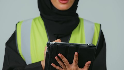 المتابعة والإشراف على العمل عبر الجهاز اللوحي , بورتريه  لمهندسة عربية خليجية سعودية ترتدي الحجاب والعباءة السوداء وسترة الحماية تمسك بيدها جهاز التابلت ، التطور والتقدم التكنولوجي في مجال الصناعة والهندسة، خلفية رمادية