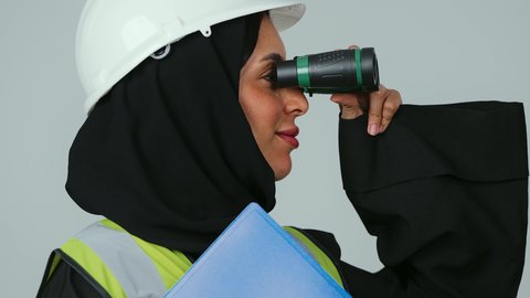 الإشراف على المشاريع، توسيع مجال الرؤية، البحث والتخطيط للمستقبل، العمل في القطاع الهندسي، بورتريه لمهندسة عربية خليجية سعودية ترتدي الحجاب والعباءة السوداء تنظر عبر منظار ثنائي العينين  تمسك بيدها اوراق عمل ، خلفية رماديه