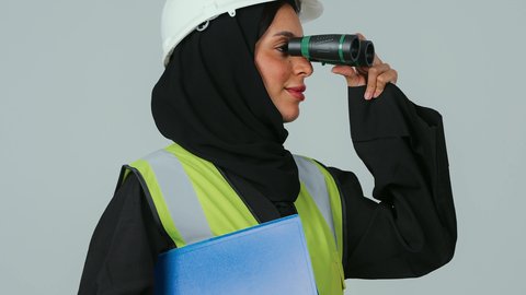 توسيع مجال الرؤية، البحث والتخطيط للمستقبل، الإشراف على المشاريع، العمل في القطاع الهندسي، بورتريه لمهندسة عربية خليجية سعودية ترتدي الحجاب والعباءة السوداء تنظر عبر منظار ثنائي العينين  تمسك بيدها اوراق عمل ، خلفية رماديه