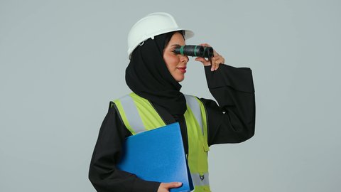 بورتريه لمهندسة عربية خليجية سعودية ترتدي الحجاب والعباءة السوداء تنظر عبر منظار ثنائي العينين  تمسك بيدها اوراق عمل ، توسيع مجال الرؤية، البحث والتخطيط للمستقبل، الإشراف على المشاريع، العمل في القطاع الهندسي، خلفية رماديه