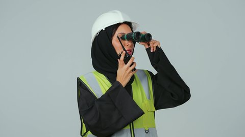 استخدام التقنيات الحديثة ، البحث والتخطيط للمستقبل، الإشراف على المشاريع، العمل في القطاع الهندسي، بورتريه لمهندسة عربية خليجية سعودية ترتدي الحجاب والعباءة السوداء تنظر عبر منظار ثنائي العينين  تقوم باستخدام جهاز الاتصال اللاسلكي، خلفية رماديه