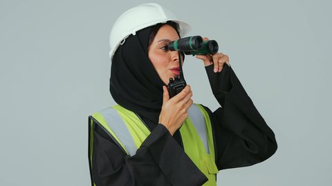 البحث والتخطيط للمستقبل، توسيع مجال الرؤية، الإشراف على المشاريع، العمل في القطاع الهندسي، بورتريه لمهندسة عربية خليجية سعودية ترتدي الحجاب والعباءة السوداء تنظر عبر منظار ثنائي العينين  تقوم باستخدام جهاز الاتصال اللاسلكي، خلفية رماديه