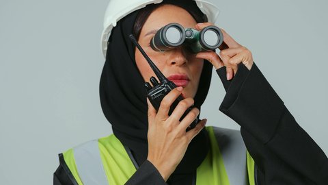 توسيع مجال الرؤية، البحث والتخطيط للمستقبل، الإشراف على المشاريع، العمل في القطاع الهندسي، بورتريه لمهندسة عربية خليجية سعودية ترتدي الحجاب والعباءة السوداء تنظر عبر منظار ثنائي العينين  تقوم باستخدام جهاز الاتصال اللاسلكي، خلفية رماديه