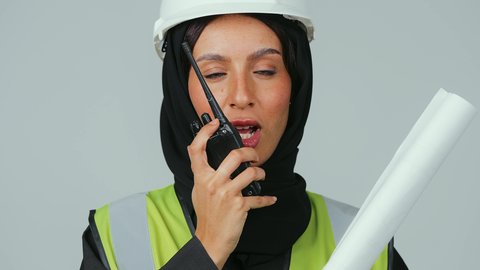 الإشراف على المشاريع، متابعة سير الاعمال، العمل في القطاع الهندسي، بورتريه لمهندسة عربية خليجية عمانية ترتدي الحجاب والعباءة السوداء وخوذة وسترة الحماية تقوم باستخدام جهاز الاتصال اللاسلكي تمسك بيدها مخطط المشروع ، خلفية رمادية