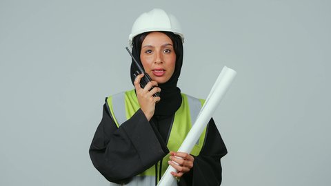 متابعة سير الاعمال، العمل في القطاع الهندسي، الإشراف على المشاريع، بورتريه لمهندسة عربية خليجية عمانية ترتدي الحجاب والعباءة السوداء وخوذة وسترة الحماية تقوم باستخدام جهاز الاتصال اللاسلكي تمسك بيدها مخطط المشروع ، خلفية رمادية