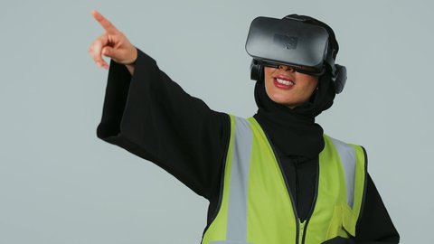 A portrait of an Arab Gulf Saudi engineer wearing a hijab and a black abaya, along with a protective vest, wearing virtual reality glasses and clicking on an item, with a gray background.