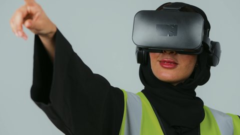 التفاعل مع الهندسة الافتراضية، تطور المجالات الهندسية عبر نظارات الفي آر، رؤية مستقبلية سعودية 2030، بورتريه  لمهندسة عربية خليجية سعودية ترتدي الحجاب والعباءة السوداء وسترة الحماية ترتدي نظارة الواقع الافتراضي تقوم بالنقر على عنصر ما، خلفية رمادية