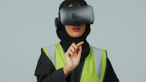A portrait of a Gulf Arab Saudi engineer wearing a hijab and a black abaya, along with a protective vest, wearing virtual reality glasses and clicking on an item. The evolution of engineering fields through VR glasses, a Saudi future vision 2030, interaction with virtual engineering, gray background.