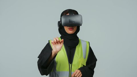 تطور المجالات الهندسية عبر نظارات الفي آر، رؤية مستقبلية سعودية 2030، التفاعل مع الهندسة الافتراضية، بورتريه  لمهندسة عربية خليجية سعودية ترتدي الحجاب والعباءة السوداء وسترة الحماية ترتدي نظارة الواقع الافتراضي تقوم بالنقر على عنصر ما، صورة لكامل الجسم، خلفية رمادية