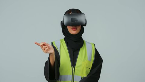 التفاعل مع الهندسة الافتراضية، تطور المجالات الهندسية عبر نظارات الفي آر، رؤية مستقبلية سعودية 2030،بورتريه  لمهندسة عربية خليجية سعودية ترتدي الحجاب والعباءة السوداء وسترة الحماية ترتدي نظارة الواقع الافتراضي تقوم بالنقر على عنصر ما، صورة لكامل الجسم، خلفية رمادية