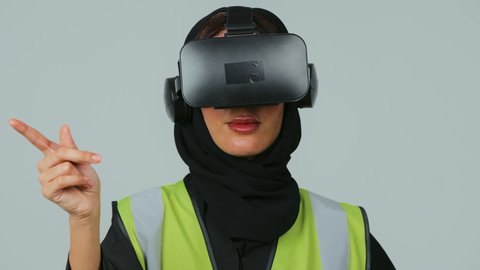 رؤية مستقبلية سعودية 2030، تطور المجالات الهندسية عبر نظارات الفي آر، التفاعل مع الهندسة الافتراضية، بورتريه  لمهندسة عربية خليجية سعودية ترتدي الحجاب والعباءة السوداء وسترة الحماية ترتدي نظارة الواقع الافتراضي تقوم بالنقر على عنصر ما، صورة لكامل الجسم، خلفية رمادية