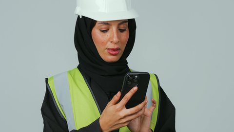العمل في القطاع الهندسي, مهن ووظائف نسائية ،بورتريه  لمهندسة عربية خليجية سعودية ترتدي الحجاب والعباءة السوداء والخوذة وسترة الحماية  تحمل الهاتف المحمول، استخدام الأجهزة التقنية الحديثة، متابعة العمل عن بعد، خلفية رماديه