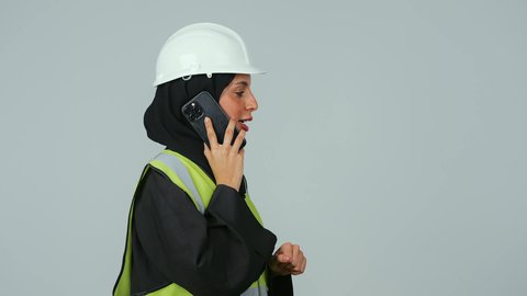 دمج التكنولوجيا المتطورة فى العمل  , بورتريه  لمهندسة عربية خليجية سعودية ترتدي الحجاب والعباءة السوداء والخوذة وسترة الحماية  تحمل الهاتف المحمول، استخدام الأجهزة التقنية الحديثة، متابعة العمل عن بعد، خلفية رماديه