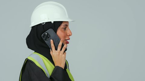 استخدام الأجهزة التقنية الحديثة،  مهن ووظائف نسائية ، العمل في القطاع الهندسي, بورتريه  لمهندسة عربية خليجية سعودية ترتدي الحجاب والعباءة السوداء والخوذة وسترة الحماية  تحمل الهاتف المحمول، متابعة العمل عن بعد، خلفية رماديه