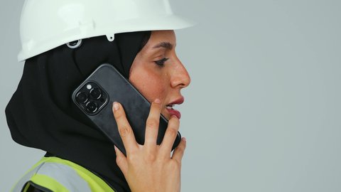 متابعة العمل عن بعد، مهن ووظائف نسائية ، العمل في القطاع الهندسي, بورتريه  لمهندسة عربية خليجية سعودية ترتدي الحجاب والعباءة السوداء والخوذة وسترة الحماية  تحمل الهاتف المحمول، استخدام الأجهزة التقنية الحديثة،  خلفية رماديه