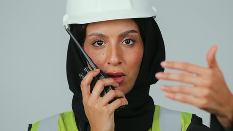 التقدم بسواعد ابناء الوطن، العمل في القطاع الهندسي، الإشراف على المشاريع، بورتريه لمهندسة عربية خليجية عمانية ترتدي الحجاب والعباءة السوداء وسترة وخوذة الحماية تقوم باستخدام جهاز الاتصال اللاسلكي، خلفية رمادية