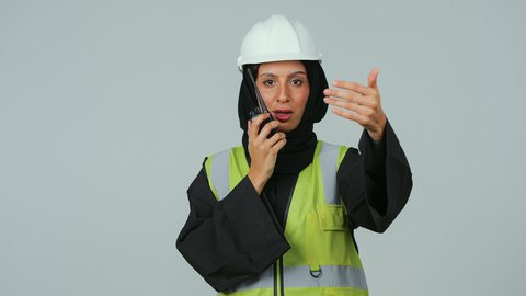 بورتريه لمهندسة عربية خليجية عمانية ترتدي الحجاب والعباءة السوداء وسترة وخوذة الحماية تقوم باستخدام جهاز الاتصال اللاسلكي، متابعة سير الاعمال، العمل في القطاع الهندسي، الإشراف على المشاريع،خلفية رمادية