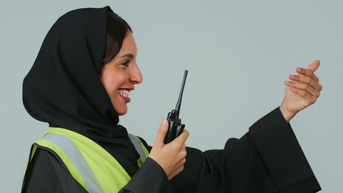 العمل في القطاع الهندسي، متابعة سير الاعمال،الإشراف على المشاريع، بورتريه لمهندسة عربية خليجية عمانية ترتدي الحجاب والعباءة السوداء وسترة الحماية تقوم باستخدام جهاز الاتصال اللاسلكي، خلفية رمادية