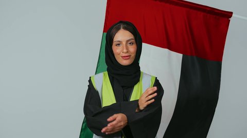 الوقوف بايدي متقاطعة ، الاعتزاز بالراية العمانية، المشاركة بالاحتفال بذكرى اليوم الوطني، إيماءات الفخر بالانتماء للبلاد، بورتريه لمهندسة عربية خليجية عمانية ترتدي الحجاب والعباءة السوداء وسترة الحماية ، خلفية علم سلطنة عمان