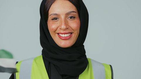 Pride in the Omani flag, participation in the celebration of National Day, gestures of pride in belonging to the country, a portrait of an Arab Gulf Omani engineer wearing a hijab and a black abaya and a protective vest holding the Sultanate's flag in her hand, with a gray background.