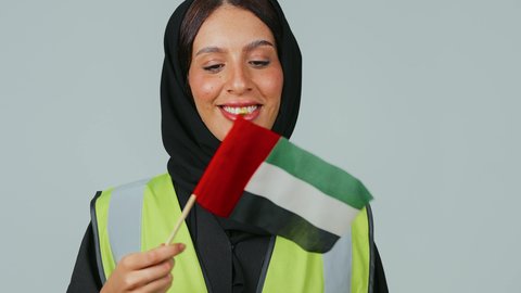 المشاركة بالاحتفال بذكرى اليوم الوطني، الاعتزاز بالراية العمانية، إيماءات الفخر بالانتماء للبلاد، بورتريه لمهندسة عربية خليجية عمانية ترتدي الحجاب والعباءة السوداء وسترة الحماية تحمل بيدها علم السلطنة ، خلفية رمادية