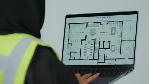 بورتريه لمهندسة عربية خليجية سعودية ترتدي الحجاب والعباءة السوداء تقوم بالتعديل على مخطط الإعمار عن طريق التطبيقات والمواقع الهندسية, إدارة ومتابعة العمل، دمج التكنولوجيا الحديثة في القطاع الهندسي, متابعة المشاريع الإنشائية عبر الحاسوب المحمول, خلفية رمادية