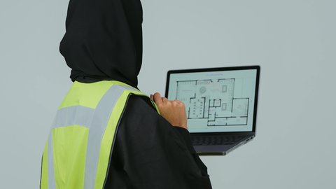 متابعة المشاريع الإنشائية عبر الحاسوب المحمول, بورتريه لمهندسة عربية خليجية سعودية ترتدي الحجاب والعباءة السوداء تقوم بالتعديل على مخطط الإعمار عن طريق التطبيقات والمواقع الهندسية, إدارة ومتابعة العمل، دمج التكنولوجيا الحديثة في القطاع الهندسي, خلفية رمادية