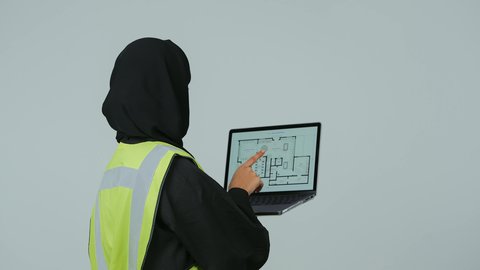 دمج التكنولوجيا الحديثة في القطاع الهندسي, متابعة المشاريع الإنشائية عبر الحاسوب المحمول, بورتريه لمهندسة عربية خليجية سعودية ترتدي الحجاب والعباءة السوداء تقوم بالتعديل على مخطط الإعمار عن طريق التطبيقات والمواقع الهندسية, إدارة ومتابعة العمل، خلفية رمادية