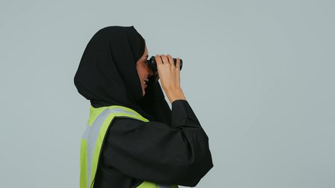 الإشراف على المشاريع، توسيع مجال الرؤية، البحث والتخطيط للمستقبل، العمل في القطاع الهندسي، بورتريه لمهندسة عربية خليجية سعودية ترتدي الحجاب والعباءة السوداء وسترة الحماية تنظر عبر منظار ثنائي العينين، خلفية رماديه