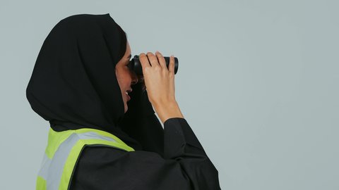 البحث والتخطيط للمستقبل، توسيع مجال الرؤية،الإشراف على المشاريع، العمل في القطاع الهندسي، بورتريه لمهندسة عربية خليجية سعودية ترتدي الحجاب والعباءة السوداء وسترة الحماية تنظر عبر منظار ثنائي العينين، خلفية رماديه