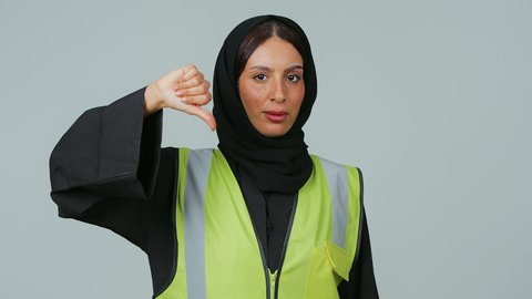 بورتريه لمهندسة عربية خليجية سعودية ترتدي الحجاب والعباءة السوداء تخفض اصبع الابهام بايماءات الانزعاج, الشعور بالرفض والامتناع, تقدم وتطور السعودية على يد أبناء الوطن، مفهوم الهندسة وأعمال البناء, خلفية رمادية