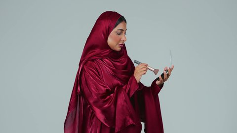 الاهتمام والعناية بالبشرة والجمال, الشغف بوضع المكياج,  بورتريه لسيدة عربية خليجية سعودية سمراء ترتدي العباءة والحجاب تمسك بيديها علبة وفرشاة أحمر الخدود, النظر إلى الكاميرا بإيماءات السعادة والسرور, مفهوم التزين والتجمل, خلفية رمادية