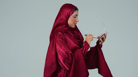 مفهوم الانوثة والنعومة , الاهتمام والعناية بالبشرة والجمال, الشغف بوضع المكياج,  بورتريه لسيدة عربية خليجية سعودية سمراء ترتدي العباءة والحجاب تمسك بيديها علبة وفرشاة أحمر الخدود, النظر إلى الكاميرا بإيماءات السعادة والسرور, خلفية رمادية