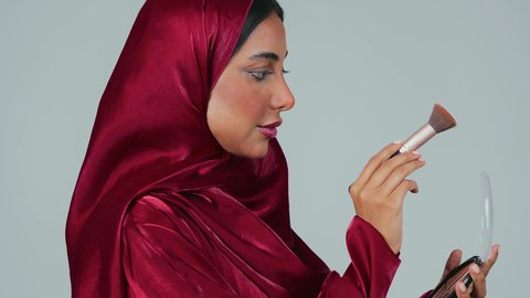 مفهوم التزين والتجمل, الاهتمام والعناية بالبشرة والجمال, الشغف بوضع المكياج,  بورتريه لسيدة عربية خليجية سعودية سمراء ترتدي العباءة والحجاب تمسك بيديها علبة وفرشاة أحمر الخدود, النظر إلى الكاميرا بإيماءات السعادة والسرور, خلفية رمادية