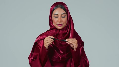 الإهتمام بالجمال والمظهر الخارجي،استخدام الماسكرا لإبراز جمال العينين، دورات تعليمية لمكياج تتوريال, خبيرة مستحضرات التجميل النسائية، بورتريه لسيدة عربية خليجية سعودية سمراء ترتدي العباءة والحجاب تقوم بوضع الميكاب، خلفيه رمادية