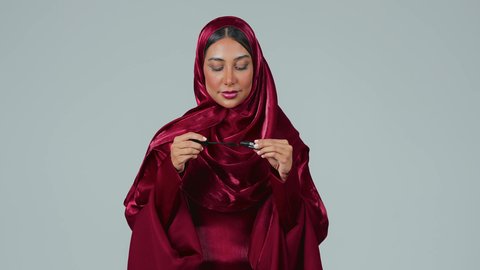 استخدام الماسكرا لإبراز جمال العينين، الإهتمام بالجمال والمظهر الخارجي، دورات تعليمية لمكياج تتوريال, خبيرة مستحضرات التجميل النسائية، بورتريه لسيدة عربية خليجية سعودية سمراء ترتدي العباءة والحجاب تقوم بوضع الميكاب، خلفيه رمادية