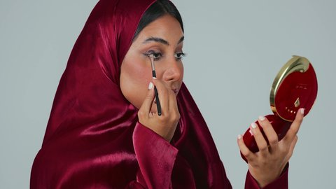 التسويق لمنتجات التجميل، أدوات مكياج تتوريال ، الإهتمام بالجمال والمظهر الخارجي ،بورتريه لامراة عربية خليجية سعودية سمراء ترتدي العباءة والحجاب تستخدم  فرشاة ظل العيون ، خلفية رمادية