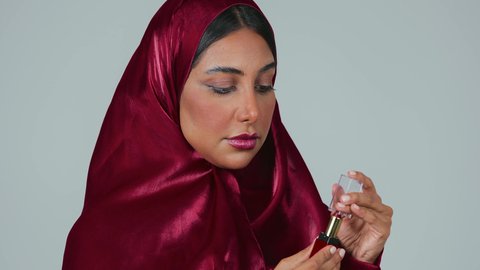 مفهوم الانوثة والنعومة ،  الإهتمام بالجمال والبشرة، ادوات مكياج تتوريال، بورتريه لمرأة عربية خليجية سعودية سمراء ترتدي العباءة والحجاب تحمل إحدى أدوات التجميل (قلم أحمر الشفاة) ، خلفية رمادية