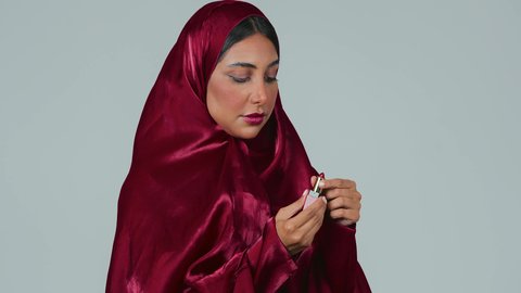ادوات مكياج تتوريال، مفهوم الإهتمام بالجمال والبشرة،بورتريه لمرأة عربية خليجية سعودية سمراء ترتدي العباءة والحجاب تحمل إحدى أدوات التجميل (قلم أحمر الشفاة) ، خلفية رمادية
