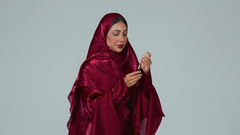 مفهوم الإهتمام بالجمال والبشرة، ادوات مكياج تتوريال، بورتريه لمرأة عربية خليجية سعودية سمراء ترتدي العباءة والحجاب تحمل إحدى أدوات التجميل (قلم أحمر الشفاة) ، خلفية رمادية